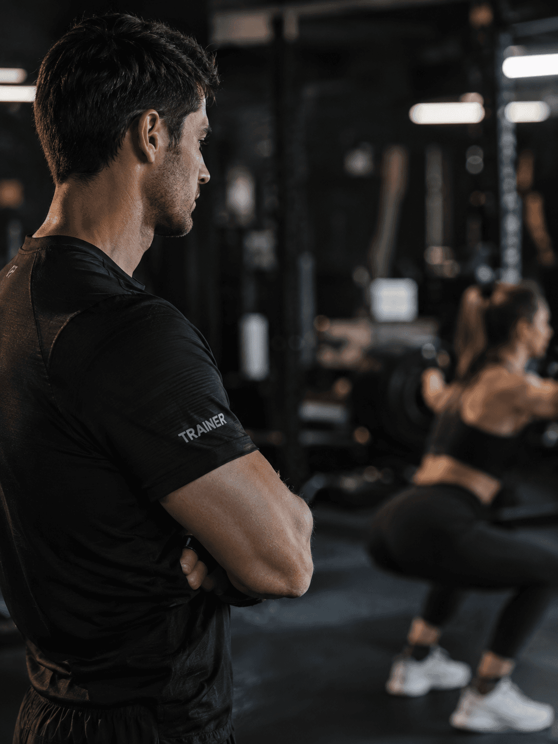 coach in profile watching a member's set, arms folded, out of focus floor behind